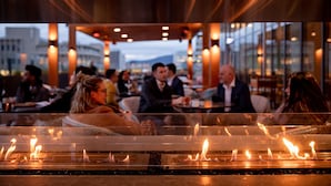 Een eethoek met tafels en stoelen, een open haard in de voorgrond en een stadszicht door grote ramen.