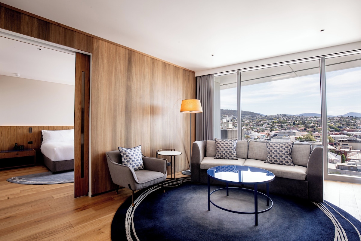 Habitación de hotel moderna con cama, sofá, una mesita y vista a un paisaje urbano a través de grandes ventanales.