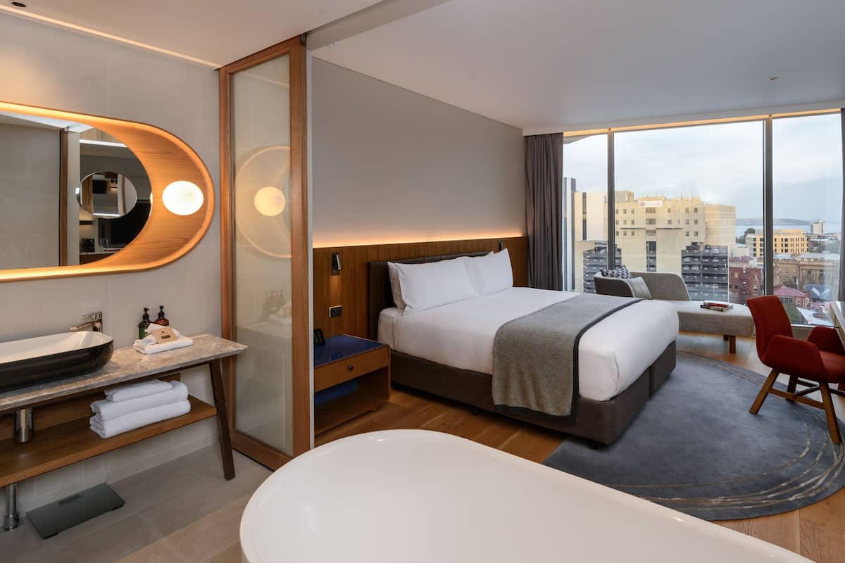 Een moderne hotelkamer met een groot bed, een afzonderlijke zithoek, een badkamer met bad en een stadszicht door grote ramen.