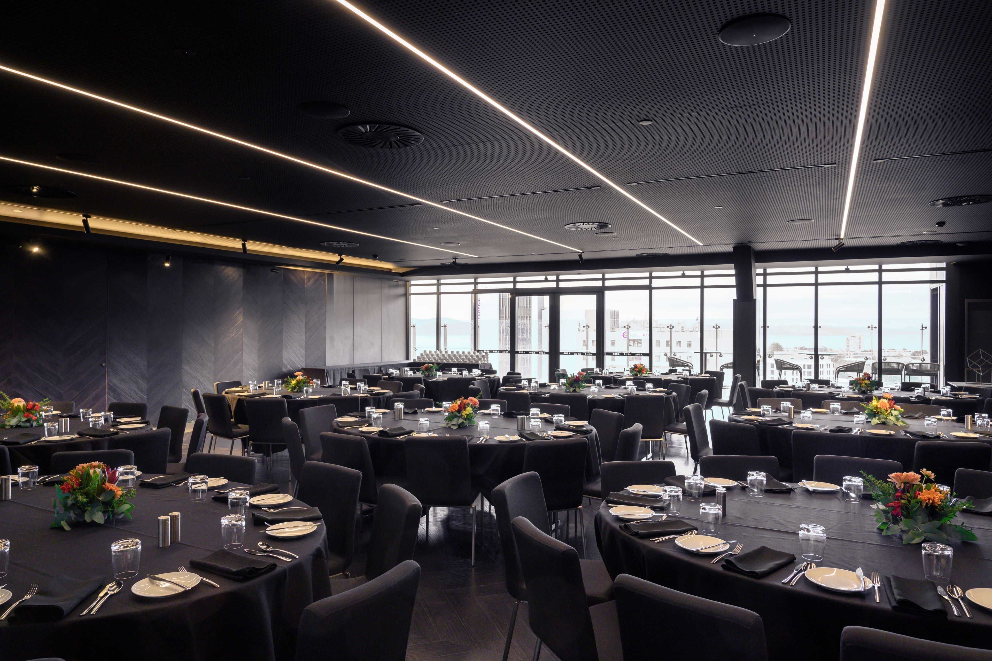 Une salle de conférence dotée de plusieurs tables dressées pour un repas, de grandes fenêtres offrant une vue sur la ville et d’un plafond au design moderne.