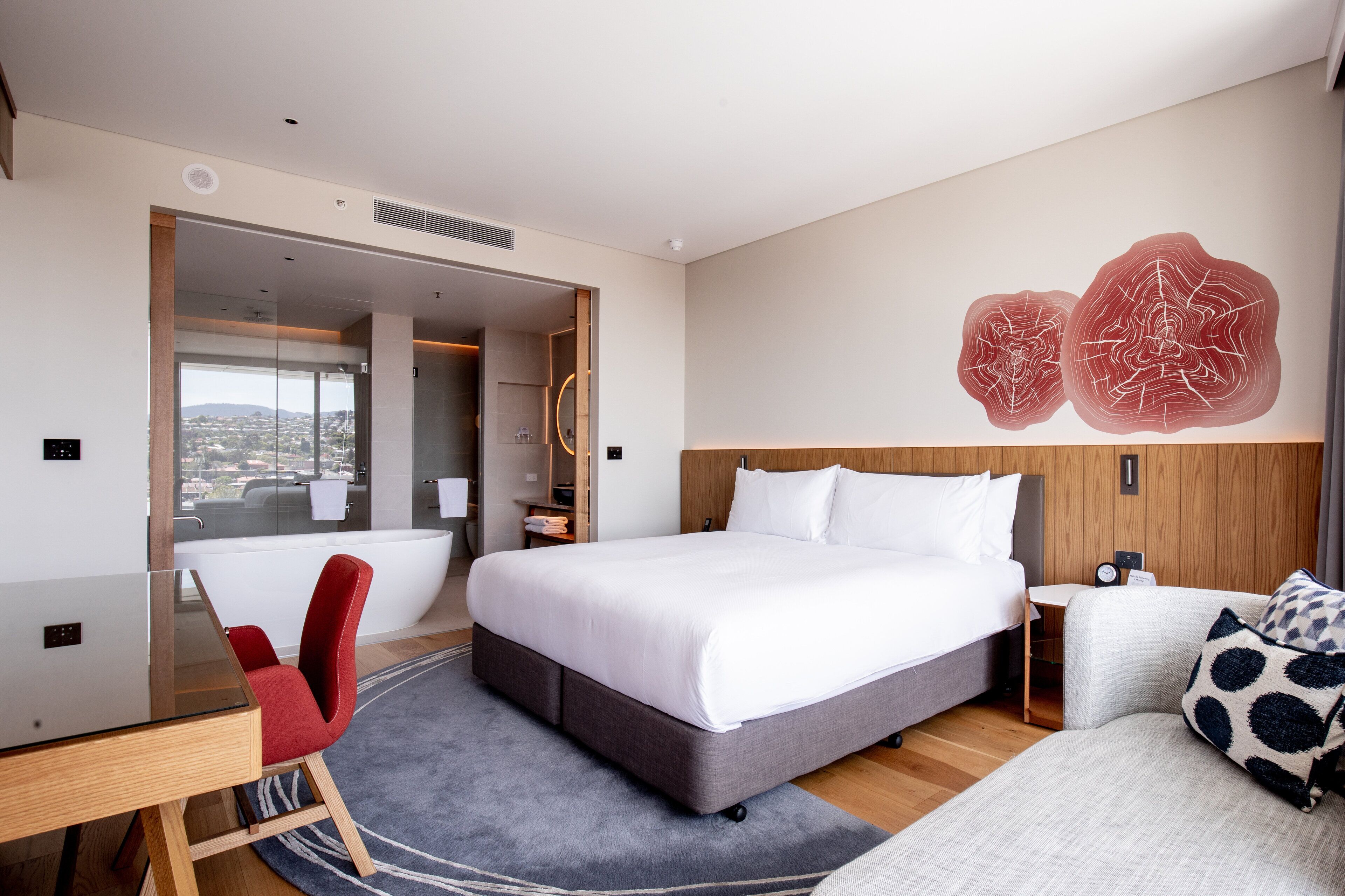 A modern hotel room with a large bed, a desk, a red chair, a gray sofa, and a view of the city through a glass door.