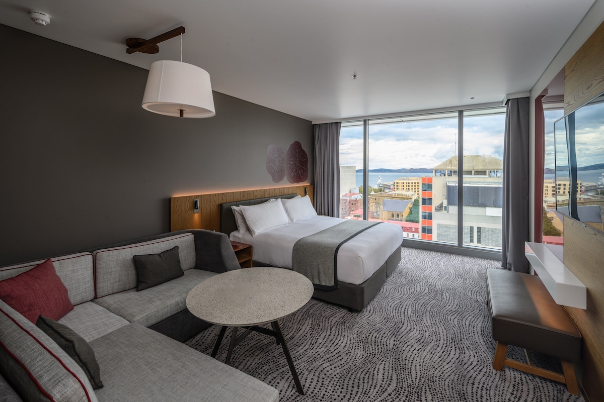 Habitación de hotel moderna con una cama grande, un sofá, una pequeña mesa redonda y vistas a la ciudad a través de amplios ventanales.