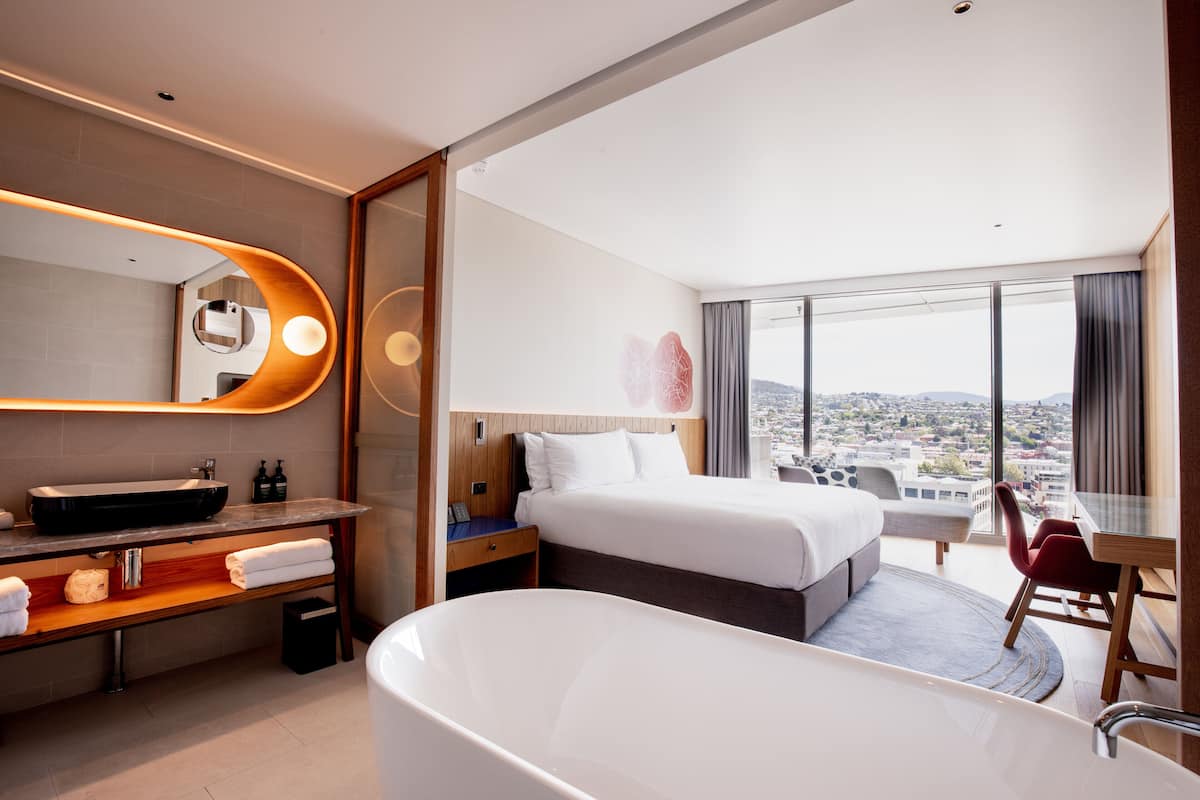 Een moderne hotelkamer met een groot bed, een afzonderlijke zithoek met een tafel en stoelen, een badkuip, een wastafel met spiegel en een stadszicht door grote ramen.