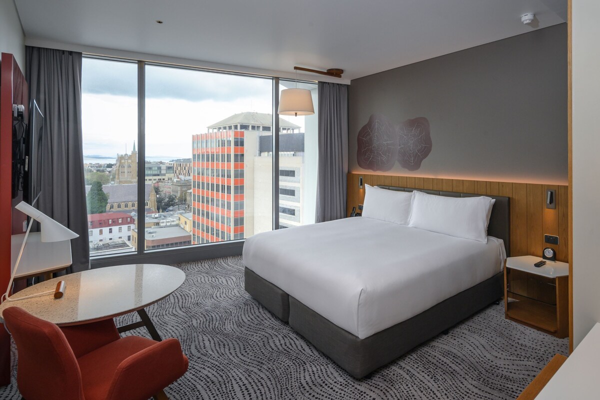 A hotel room with a large bed, a desk, a chair, a bedside table, and a view of the cityscape.