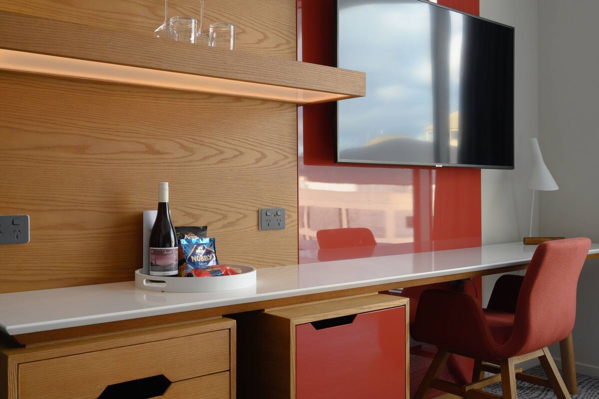 Une chambre d’hôtel moderne avec un bureau en bois, une chaise rouge, un téléviseur à écran plat et une bouteille de vin.