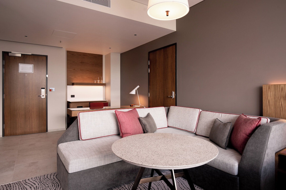 Una habitación de hotel moderna con un sofá modular gris, una mesa redonda y un mueble de madera.