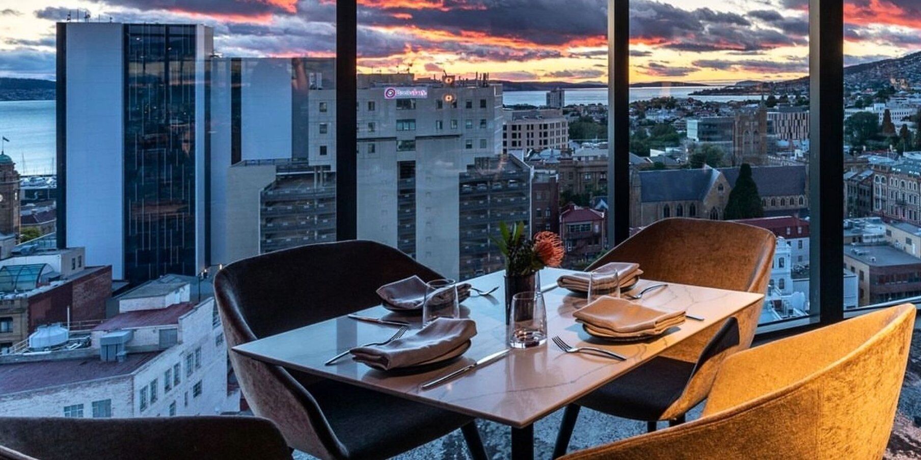 Un comedor con una mesa dispuesta para dos, un florero con una flor y vistas a la ciudad a través de amplios ventanales.