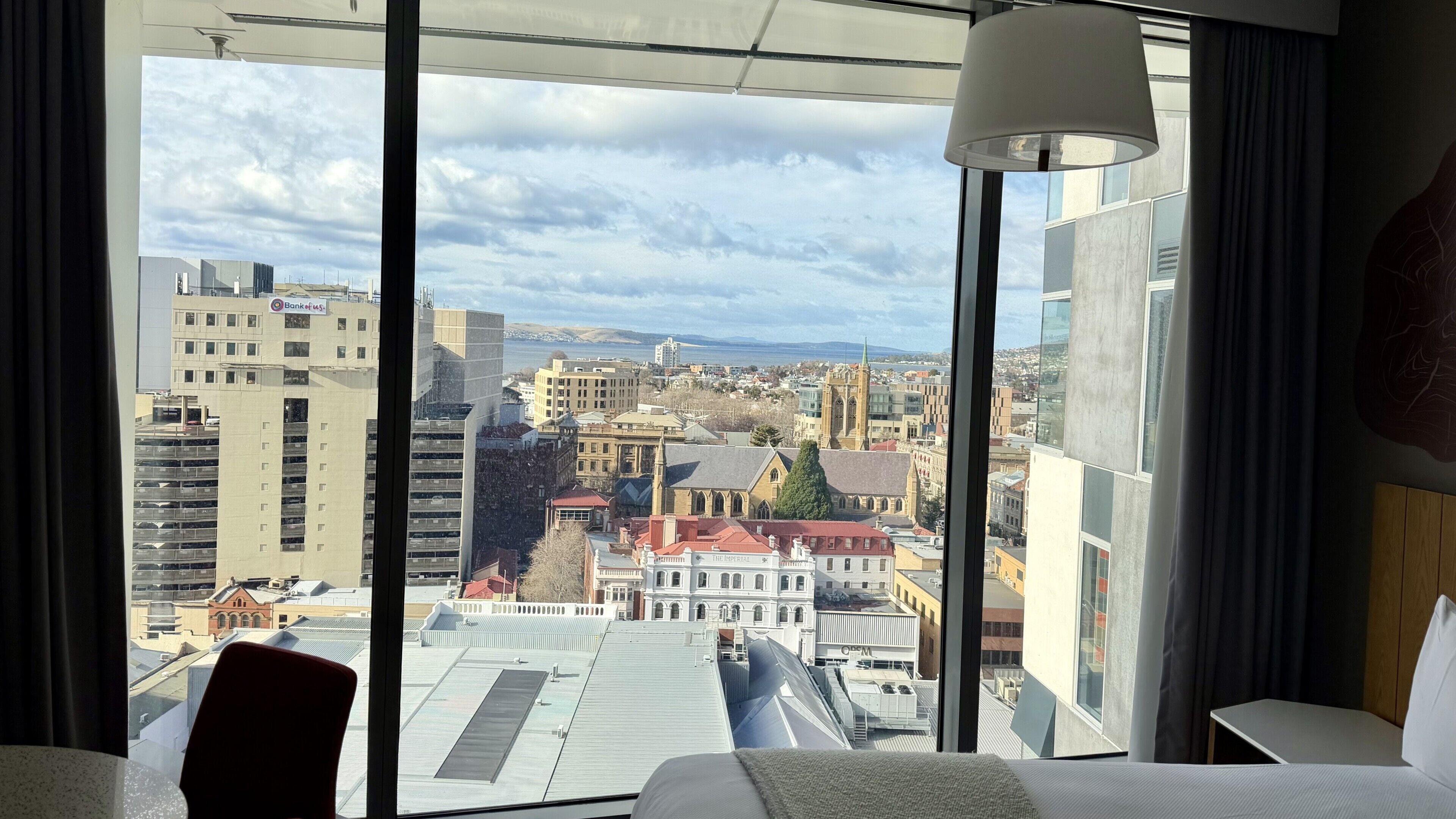 A hotel room with a large window offering a city view, a bed, a chair, and a lamp.