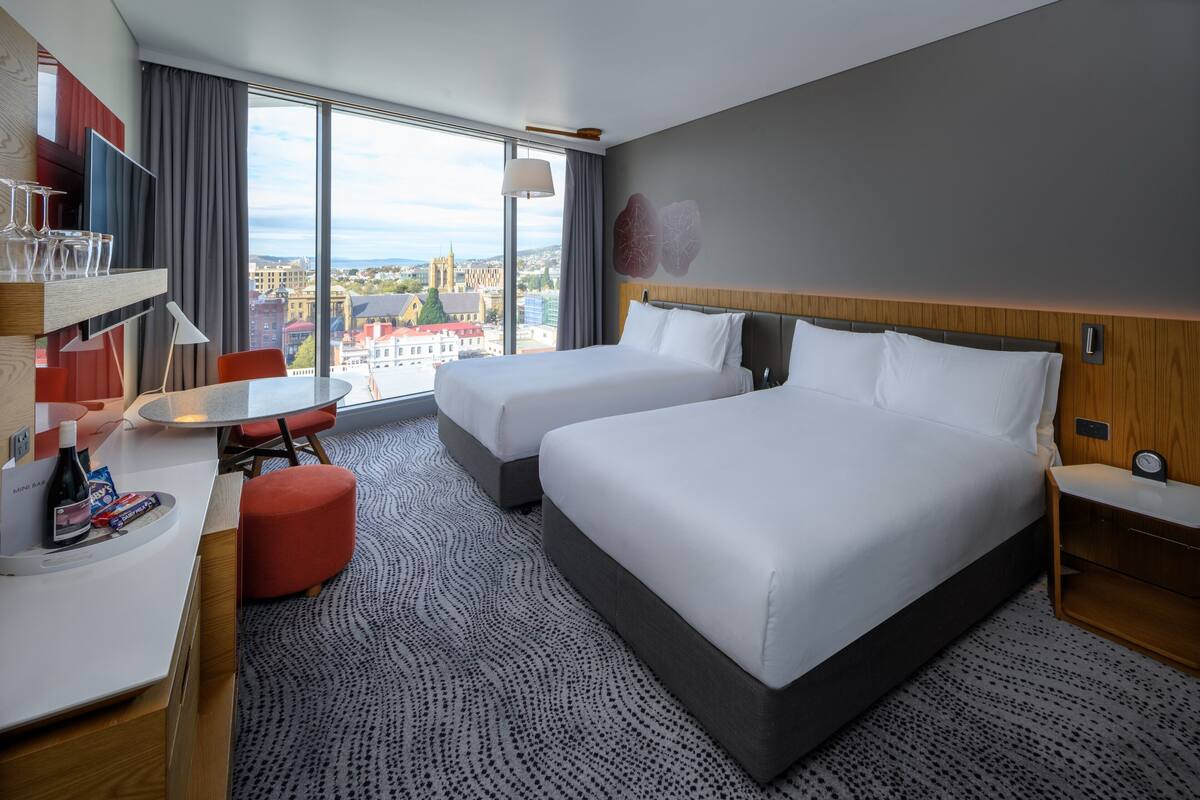Een hotelkamer met twee bedden, een bureau met stoel, een kleine tafel en een groot raam met uitzicht op de stad.
