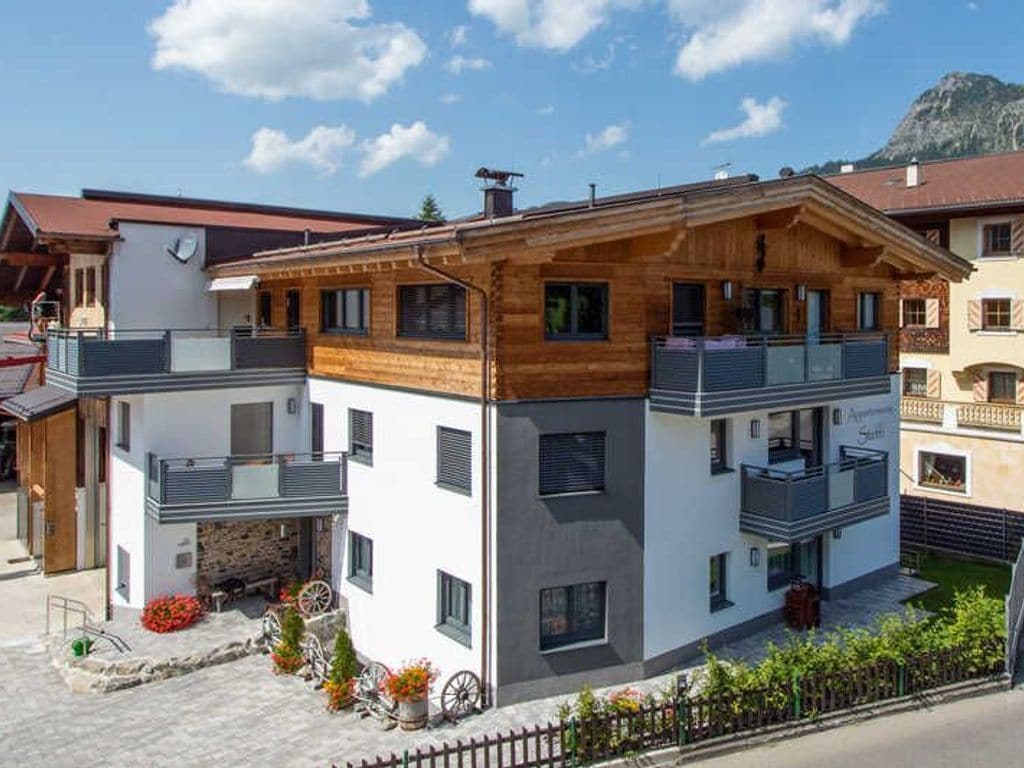 A modern two-story building with a balcony, a stone fireplace, and a small garden with flowers and a wheelbarrow.