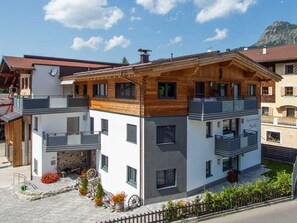 A modern two-story building with a balcony, a stone fireplace, and a small garden with flowers and a wheelbarrow.