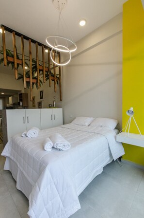 Un dormitorio moderno con una cama, ropa de cama blanca, un escritorio con lámpara y una pared de acento amarilla.