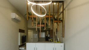 Quarto com cama, roupa de cama branca, luminária no teto, televisão e área de cozinha visível através de um espaço aberto.