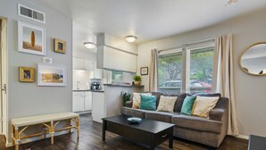 A modern living room with a sofa, coffee table, and a bench. There is a kitchen area with cabinets and a window with curtains. - Enfield #114 xxxxxxx (Austin)