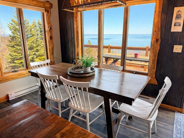 Un comedor con mesa de madera y sillas blancas, ventanas amplias con vista al mar y una terraza de madera afuera.