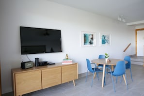 A modern living room with a flat-screen TV mounted on the wall, a wooden cabinet, a round table with blue chairs, and a staircase in the background.