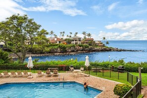 A swimming pool with lounge chairs and umbrellas, overlooking a rocky coastline with houses and palm trees.