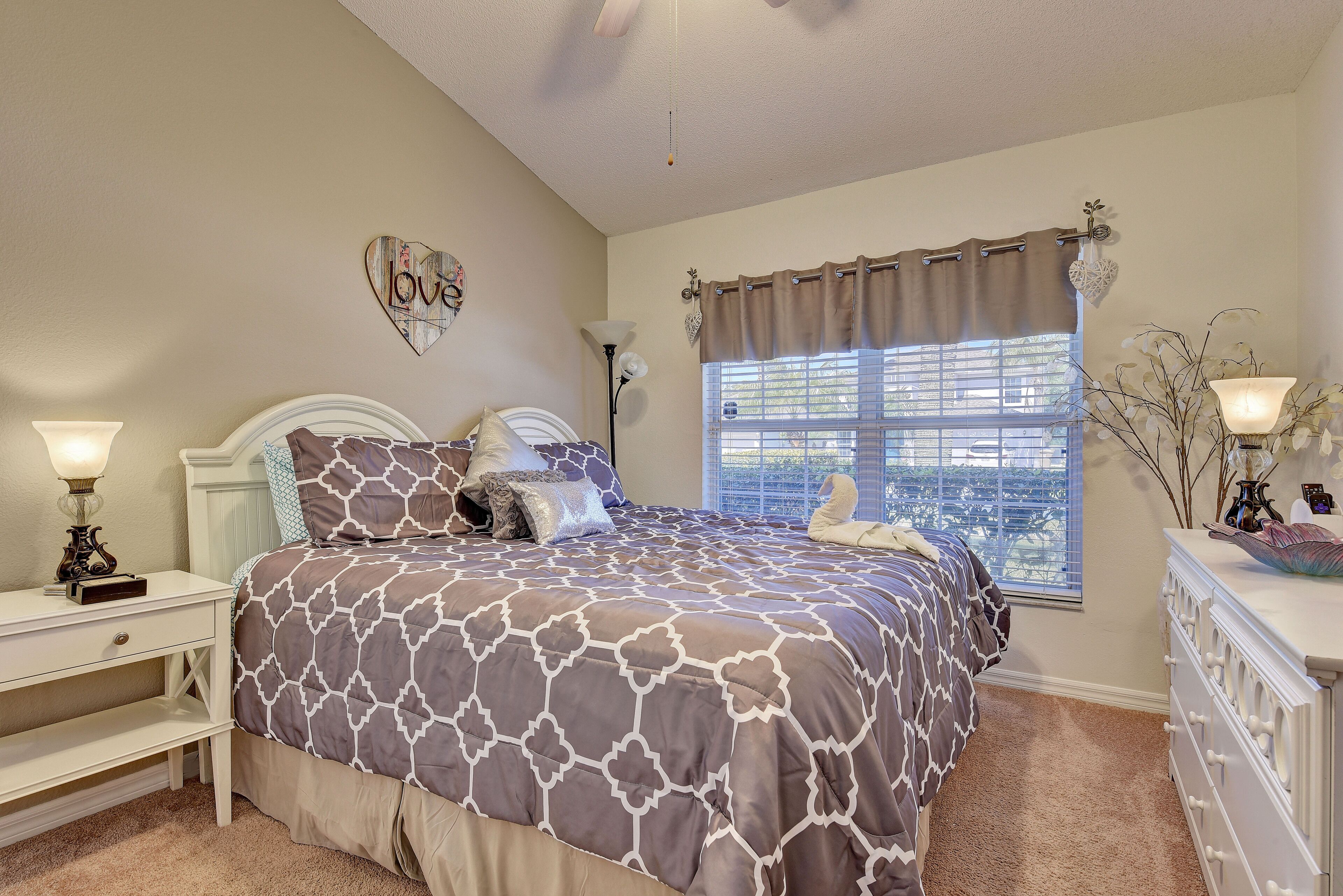 A bedroom with a bed, a nightstand, a dresser, a window with blinds, and a wall decoration.