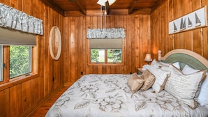 A wooden-paneled bedroom with a bed, a ceiling fan, a window with blinds, and a wall-mounted mirror.