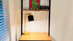 A corner desk with a wooden top, a black lamp, and a chair. There is a shelf above the desk with a framed picture and a book titled "BLACKBEARD". - Sosuite | Arcade 2BR Apt w/ W/D (Philadelphia)