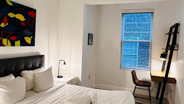 A bedroom with a large bed, a desk, a chair, a window with blinds, and a ceiling light. - Sosuite | Arcade 2BR Apt w/ W/D (Philadelphia)
