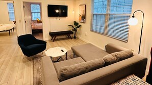 A modern living room with a sofa, a chair, a bench, and a TV. There is a window with blinds and a floor lamp. - Sosuite | Arcade 2BR Apt w/ W/D (Philadelphia)