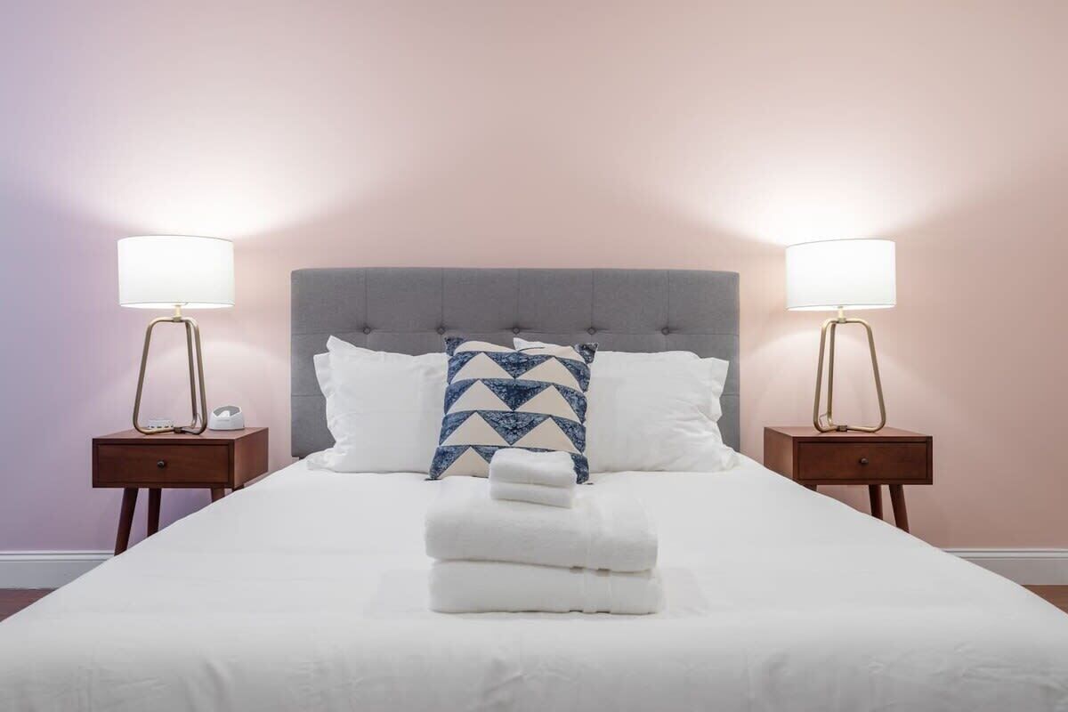 A neatly made bed with white linens, a grey upholstered headboard, and two bedside lamps on matching nightstands.