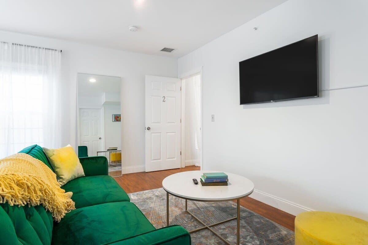A modern living room with a green sofa, a small round table, and a flat-screen TV mounted on the wall.