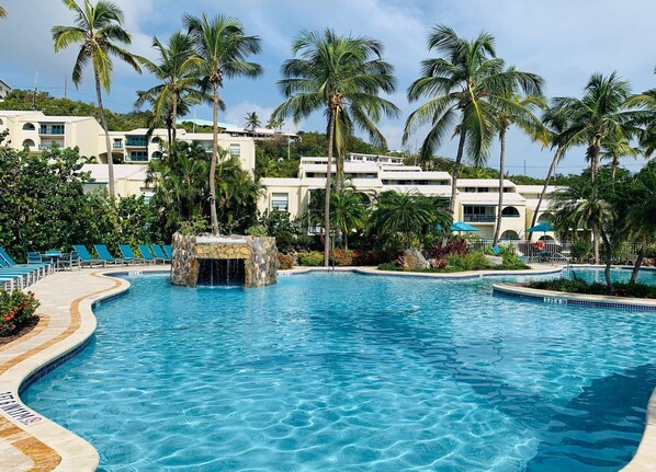 A resort pool with a stone waterfall feature, surrounded by palm trees and lounge chairs.