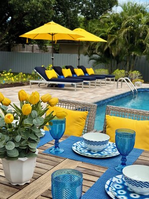 Un espace aménagé au bord de la piscine, avec des chaises longues, une table à manger dressée avec de la vaisselle bleue et un pot de fleurs contenant des fleurs jaunes.