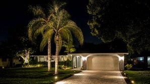 A well-lit driveway leading to a garage, surrounded by palm trees and a landscaped garden. - Modern Coastal Pool Retreat • Private Heated Pool • Minutes to Siesta Key Beach (Sarasota)