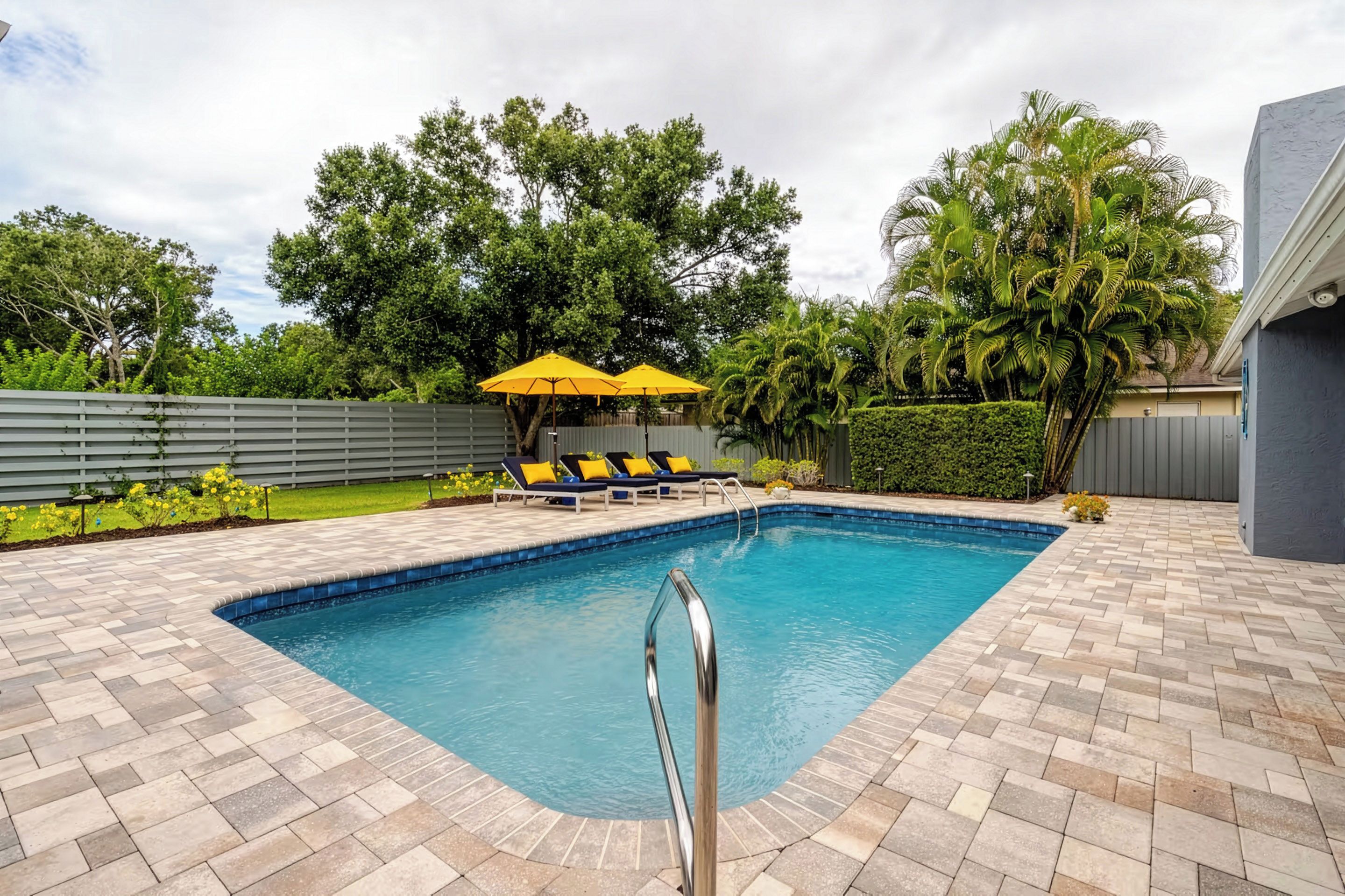 A swimming pool with a metal ladder, surrounded by a tiled deck, with lounge chairs and yellow umbrellas in the background.
