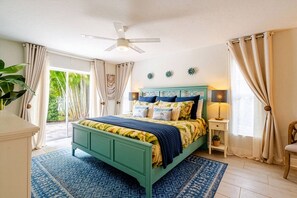 A bedroom with a bed, a ceiling fan, a nightstand, a lamp, and a view of greenery outside. - Modern Coastal Pool Retreat • Private Heated Pool • Minutes to Siesta Key Beach (Sarasota)