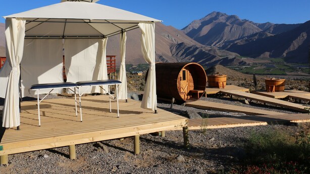 Een houten terras met een overkapping, houten vaten en een bergachtige achtergrond.