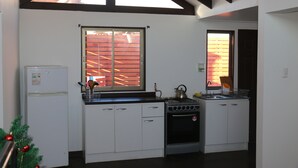 Een kleine keuken met witte kastjes, een fornuis, een koelkast en een raam met uitzicht op een bakstenen muur.