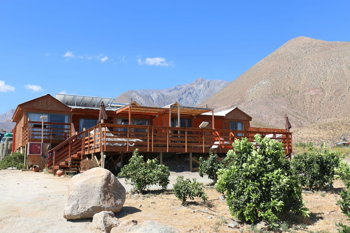 A wooden cabin with a balcony, surrounded by desert vegetation and mountains.
