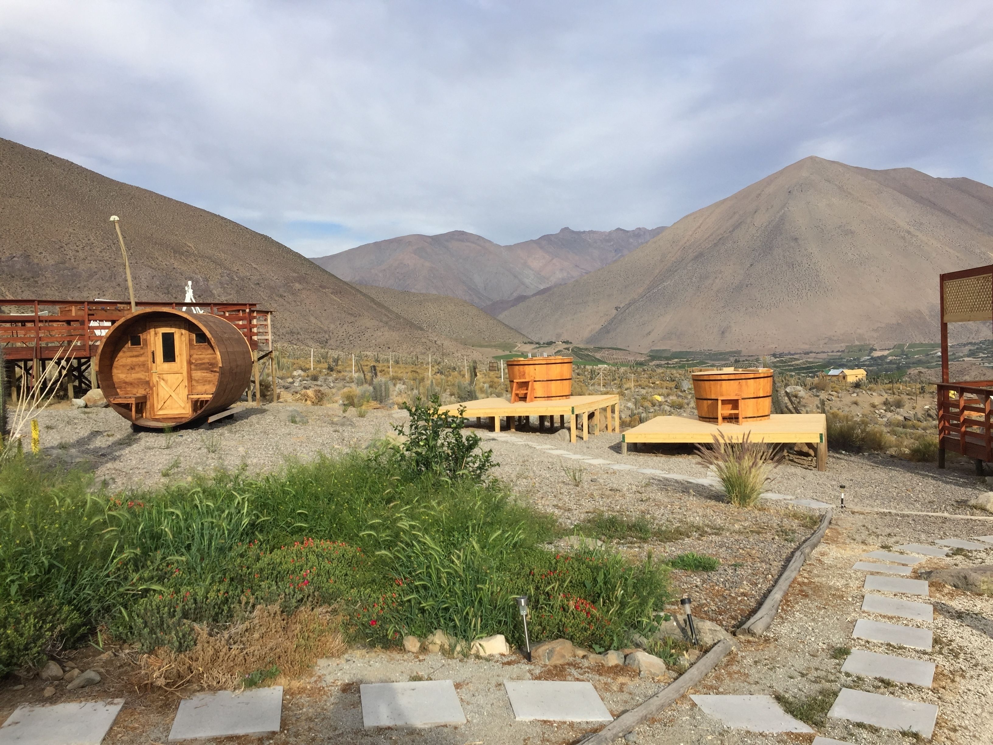 A rustic cabin with a wooden barrel structure, surrounded by a dry landscape with mountains in the background.