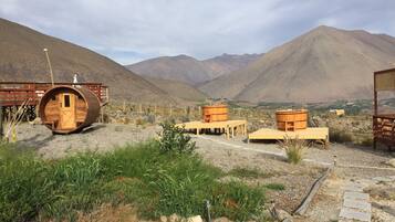 A rustic cabin with a wooden barrel structure, surrounded by a dry landscape with mountains in the background.