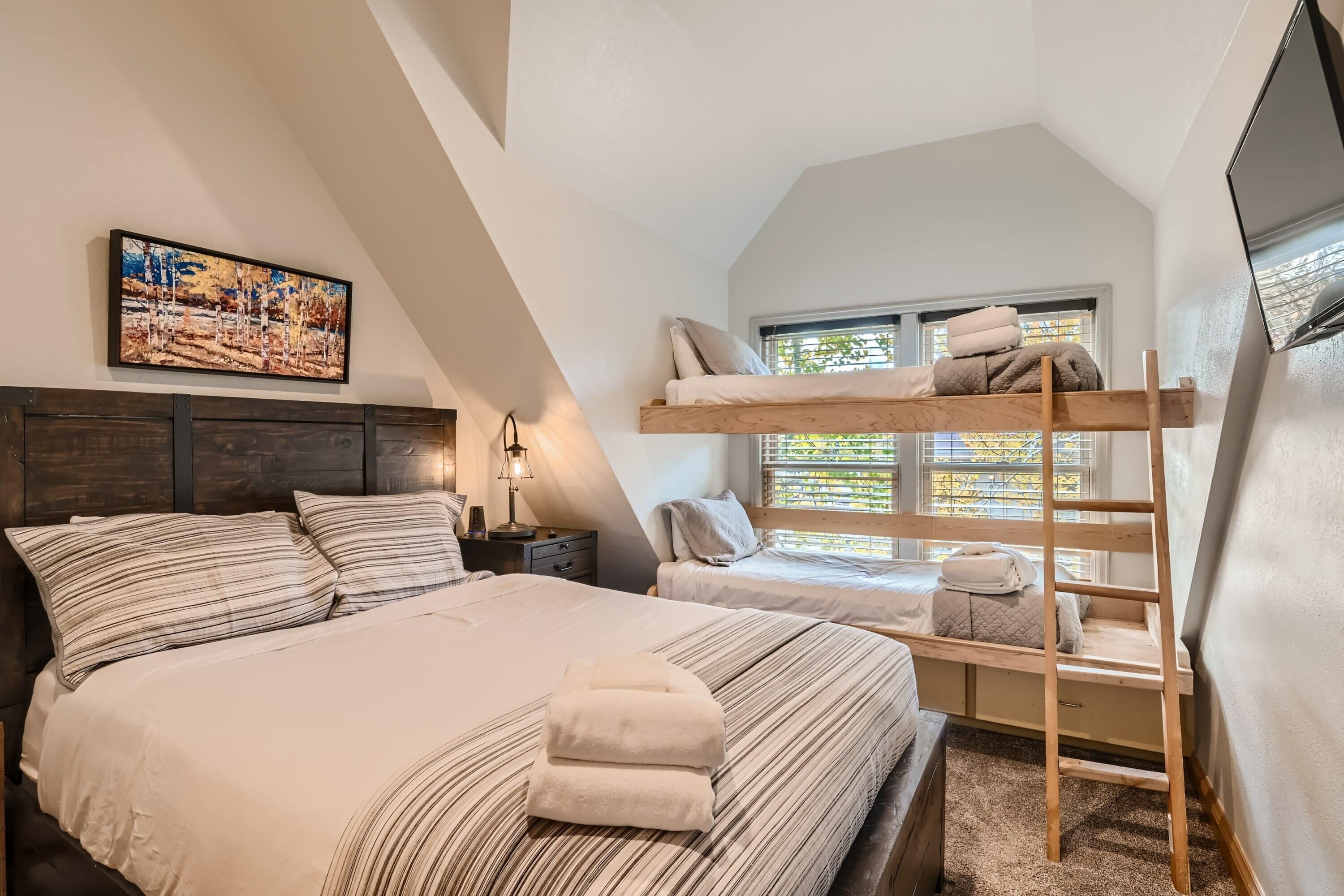 A bedroom with a bunk bed, a large bed, a ladder, a window, and a painting on the wall.
