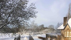 A snow-covered property with a stone wall, a paved driveway, and a building with a pitched roof.