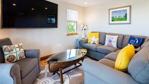 A modern living room with a large flat-screen TV, a glass coffee table, and a grey sofa with yellow and white cushions.