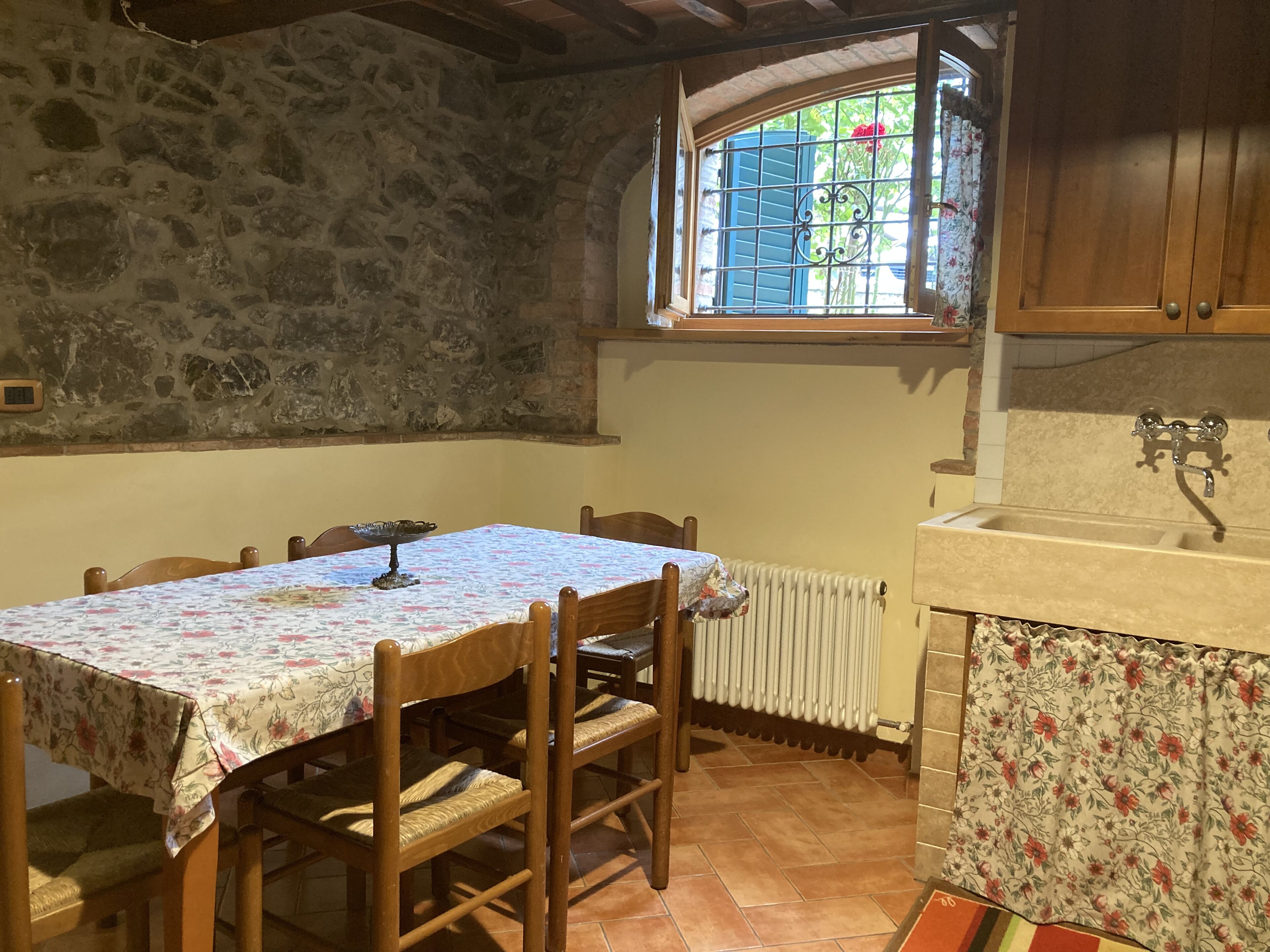 Un mur en pierre rustique, une table à manger en bois avec des chaises, une fenêtre avec des volets bleus et un coin cuisine avec un évier et un dosseret carrelé.