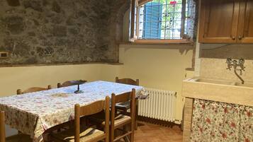 Un mur en pierre rustique, une table à manger en bois avec des chaises, une fenêtre avec des volets bleus et un coin cuisine avec un évier et un dosseret carrelé.