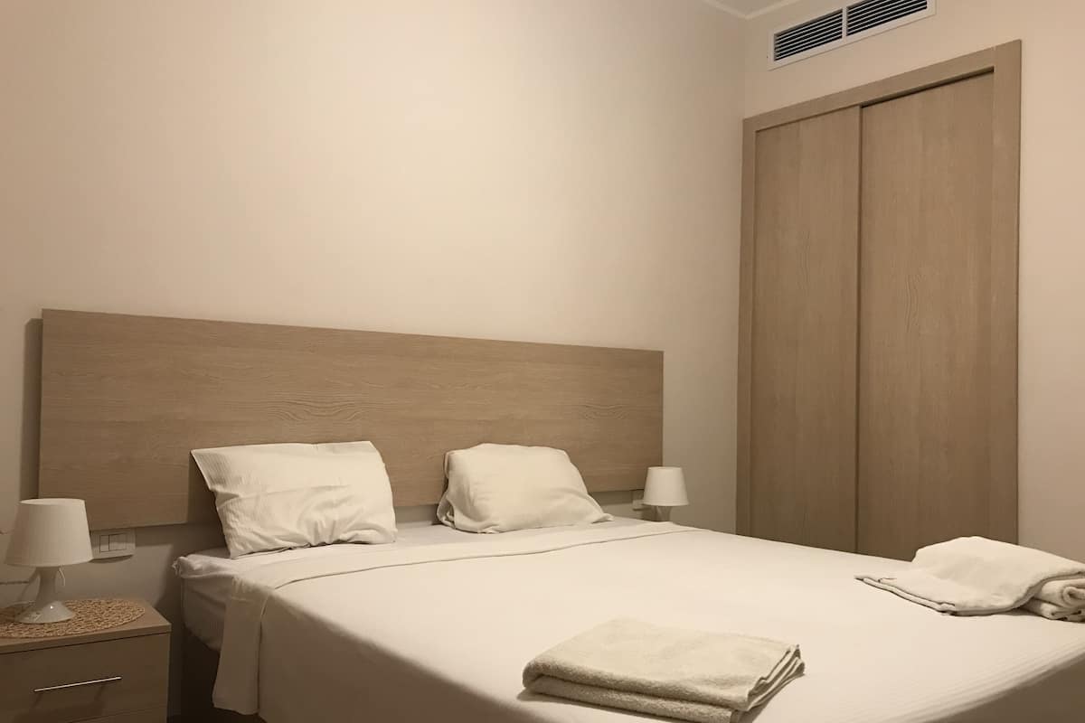 Un lit bien fait, avec du linge de lit blanc, deux oreillers et une serviette pliée posée sur le lit. Il y a une table de chevet avec une lampe et un tiroir. La chambre est dotée d’une tête de lit en bois et d’une grande armoire.