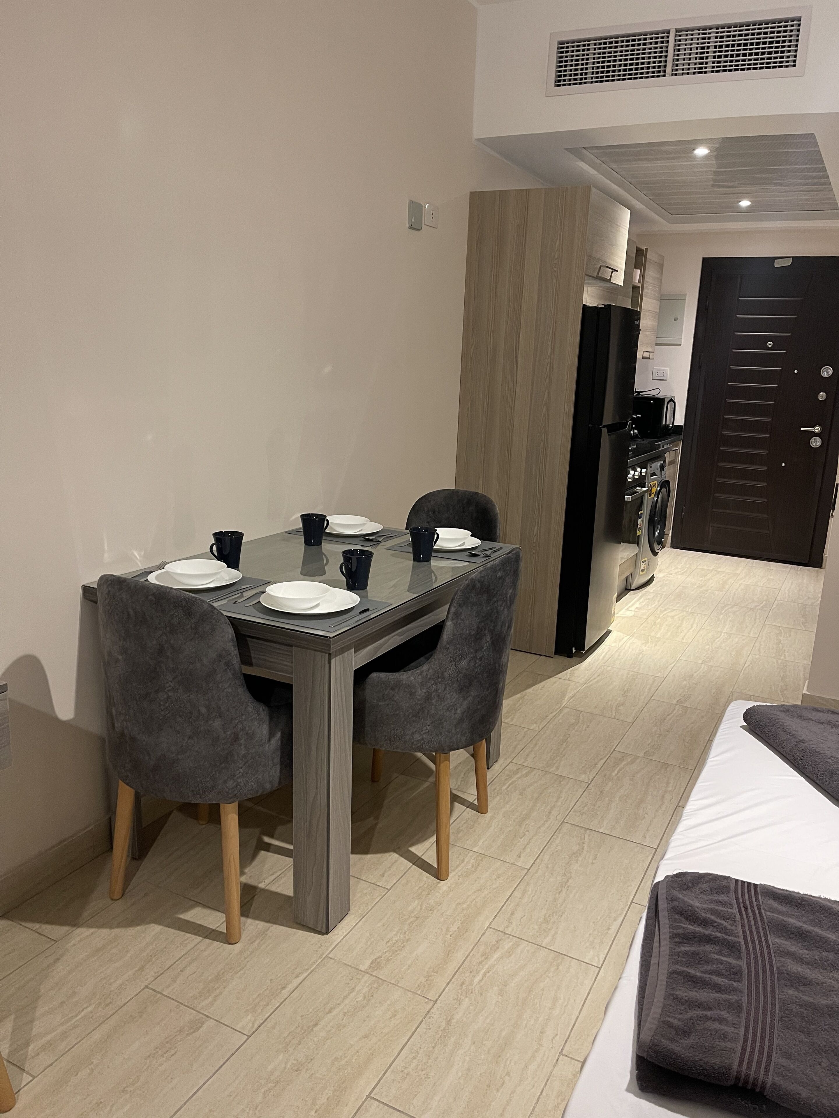 A compact dining area with a dark wooden table, four grey upholstered chairs, and a small black refrigerator in the background.