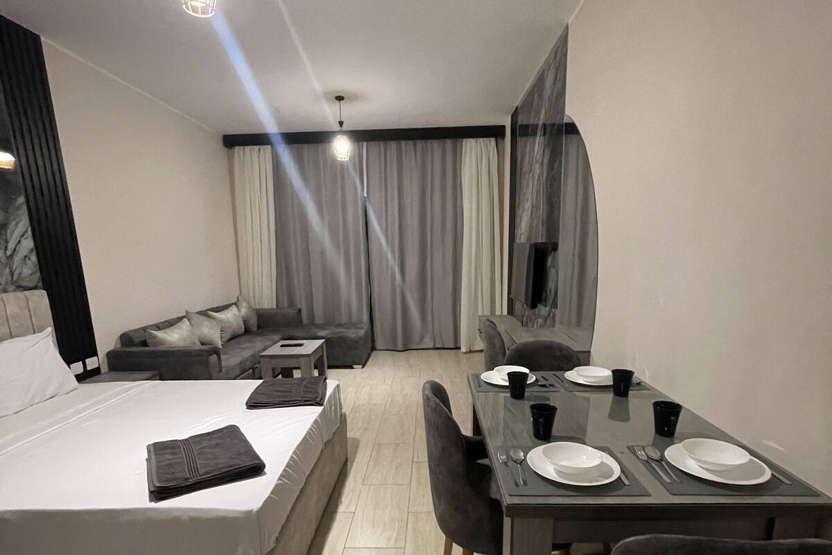 Une chambre d’hôtel moderne équipée d’un lit, d’une table à manger pour deux personnes, d’un téléviseur et d’une grande fenêtre avec des rideaux.