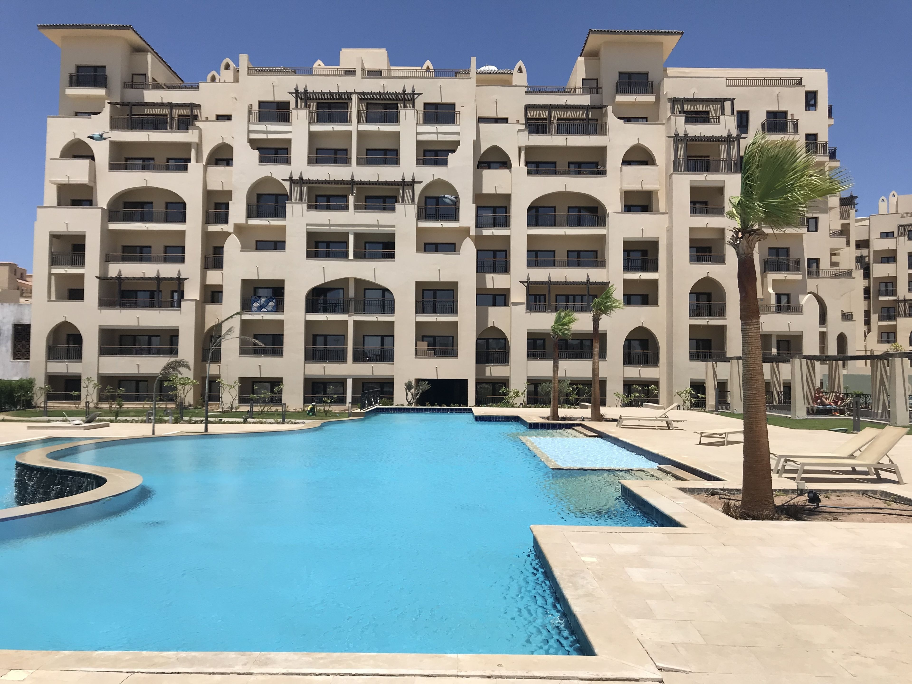A multi-story hotel with a central swimming pool, surrounded by palm trees and lounge chairs.