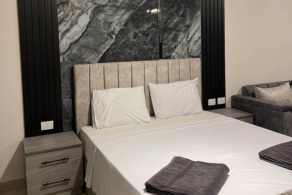 Une chambre à coucher moderne avec une tête de lit en marbre, un lit recouvert de draps blancs, deux oreillers, un canapé gris et une petite table de chevet avec des tiroirs.