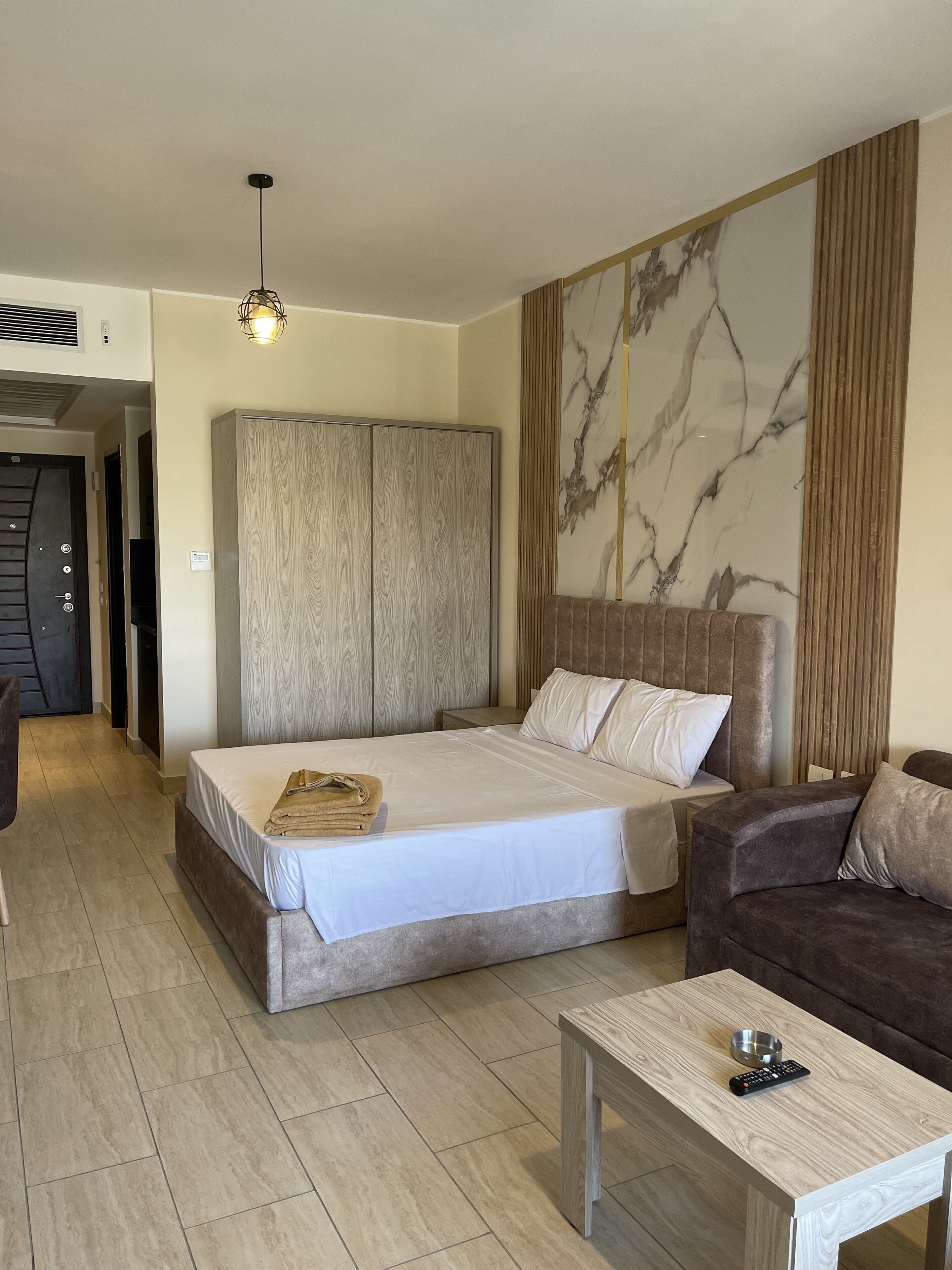 Una habitació d'hotel moderna amb un llit gran, un armari de fusta, zona de seients amb un sofà marró i una taula petita amb un comandament a distància.