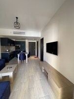 Una sala de estar moderna con un televisor de pantalla plana montado en la pared, un comedor con mesa y sillas, y una cocina con gabinetes y electrodomésticos oscuros.